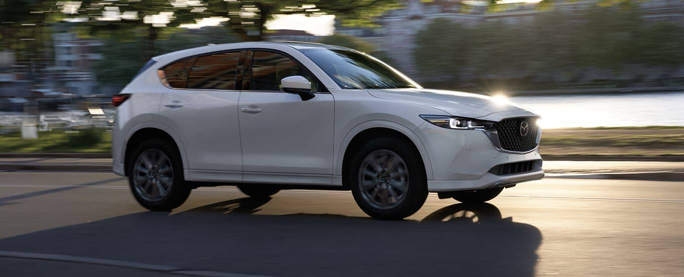 2025 White Mazda CX-5 SUV driving on a city street near water, with motion blur in the background.