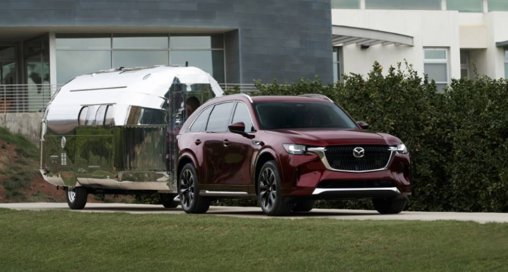 Red 2025 Mazda CX-90 SUV towing a silver Airstream trailer in front of a modern building.