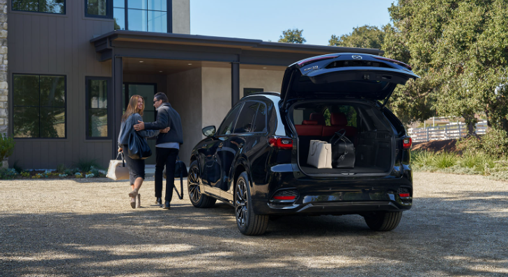 Couple walking toward a house with a black 2025 Mazda CX-70 SUV parked outside, rear hatch open showing cargo space with bags.