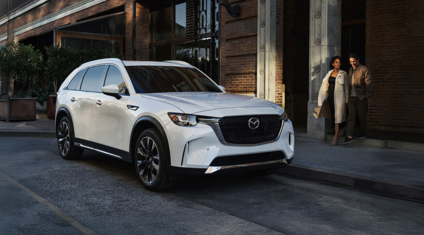 White 2026 Mazda CX-90 PHEV parked next to couple on sidewalk.